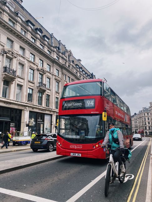A red double-decker bus displaying route 94 with the destination 'Acton Green' is moving along a city street lined with historic multi-storey buildings featuring ornate balconies and large windows. In the foreground, a cyclist wearing a dark helmet, light blue jacket, and a backpack is riding on a designated cycle lane marked by a solid white line and bicycle symbol on the asphalt. Pedestrians and parked cars are visible on the sidewalk, with some showing reflective safety vests, indicating possible use by waste collection or city staff. Overhead, there are utility wires stretching across the cloudy sky, and the overall scene captures a typical urban environment with active transportation and public bus services. The setting provides contextual relevance for independent or alternative transportation options involved in waste management or rubbish removal services contrasted against the backdrop of city life, consistent with environmentally conscious urban waste handling practices supported by local service providers such as Waste Removal Acton.
