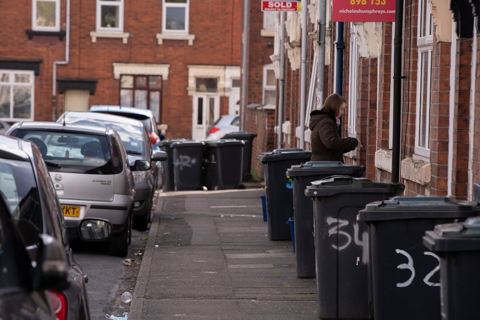 A city street scene shows a row of dark grey wheelie bins with hinged lids positioned along the sidewalk adjacent to a brick residential building. The bins are arranged in a line, some with graffiti markings in white spray paint on their sides. A woman dressed in a dark coat stands behind one of the bins, appearing to be disposing of rubbish or engaging with its contents. Behind her, the building’s exterior features multi-pane sash windows with white frames and stone lintels. The sidewalk is made of grey concrete paving slabs, with some minor debris scattered along its surface. Further along the street, parked cars in various colours are lined up, including a silver hatchback closest to the foreground, with additional vehicles visible beyond. The street and sidewalk are illuminated by ambient daylight, and in the background, a red-brick terrace with similar windows and a 'SOLD' sign on one of the properties can be seen. This scene exemplifies a typical urban setting suitable for private rubbish collection and on-site waste disposal services, as offered by Waste Removal Acton, supporting effective rubbish management for residential areas.