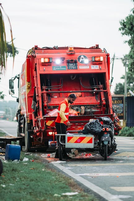 A waste collection operative wearing a high-visibility yellow and red vest is standing at the rear of a large red waste collection truck, which is parked on the side of a street. The back of the truck is open, revealing its internal mechanisms, with some debris visible on the ground near the opening. The worker appears to be handling a black plastic bag of rubbish, possibly preparing it for disposal or collection. Surrounding the scene, there is a motorbike parked close to the truck, partially blocking the street, with a small metal barrier or cart in front of the worker. In the background, a few trees, utility poles, and wires are visible, along with some distant commercial or residential buildings and a blue structure on the right side of the image. The environment indicates an urban or suburban area where private waste collection services, such as those provided by Waste Removal Acton, might operate, supporting alternative rubbish removal methods beyond local authority collection. The image is lit with natural daylight, contributing to a clear and detailed depiction of the rubbish removal process.