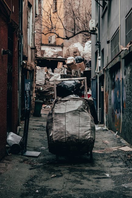 The image depicts a narrow urban alleyway cluttered with a large, wheeled garbage bag made of woven synthetic material placed in the foreground, appearing slightly overstuffed and stained. Behind it, a pile of mixed rubbish, including cardboard boxes, plastic bags, and assorted debris, is stacked haphazardly against a worn brick wall and a building with graffiti-covered metal panels. The alley is flanked by tall, closely positioned buildings with visible vents, pipes, and windows, some of which are partially obscured by leafless tree branches extending overhead. The ground is asphalt with visible dirt, grime, and scattered rubbish. The scene is lit by diffuse natural light, likely on an overcast day, creating a muted, subdued atmosphere. The setup of the waste materials suggests an instance of private rubbish clearance, possibly undertaken by a waste removal service such as Waste Removal Acton, to clear accumulated refuse from an urban environment, highlighting the necessity of independent collection efforts in densely built areas.