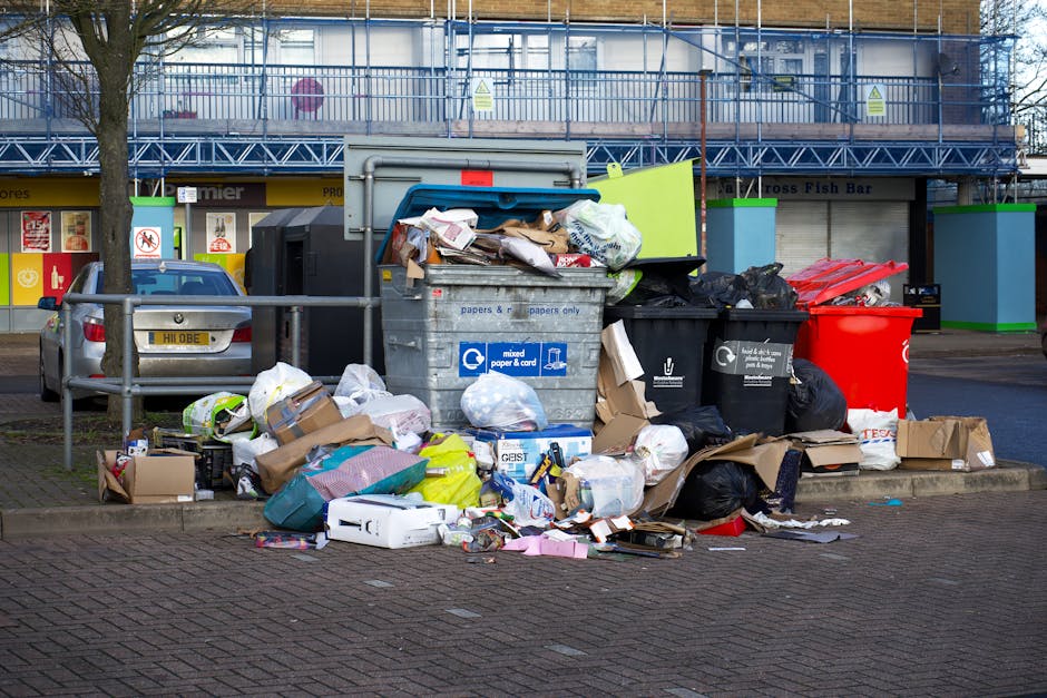 A waste collection operative wearing a high-visibility yellow and red vest is standing at the rear of a large red waste collection truck, which is parked on the side of a street. The back of the truck is open, revealing its internal mechanisms, with some debris visible on the ground near the opening. The worker appears to be handling a black plastic bag of rubbish, possibly preparing it for disposal or collection. Surrounding the scene, there is a motorbike parked close to the truck, partially blocking the street, with a small metal barrier or cart in front of the worker. In the background, a few trees, utility poles, and wires are visible, along with some distant commercial or residential buildings and a blue structure on the right side of the image. The environment indicates an urban or suburban area where private waste collection services, such as those provided by Waste Removal Acton, might operate, supporting alternative rubbish removal methods beyond local authority collection. The image is lit with natural daylight, contributing to a clear and detailed depiction of the rubbish removal process.